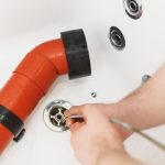 A person uses a plumbing snake to clear a clogged drain in a white sink, with an orange pipe visible to the side.