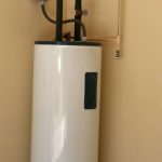 Water heater with a cylindrical white body and pipes, installed on a beige wall, by Quahog Plumbing.