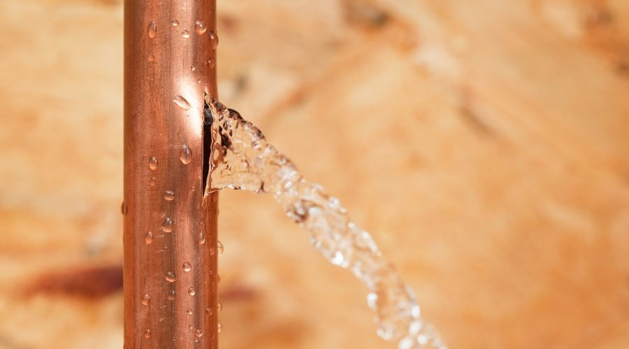 Copper pipe with water leaking from a crack, droplets on surface, highlighting plumbing issue related to water leaks.